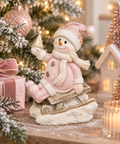 Pink snowman on sledge figurine displayed on a wooden surface beside a pink gift box and candle, with a blush Christmas tree and pink baubles in warm bokeh light