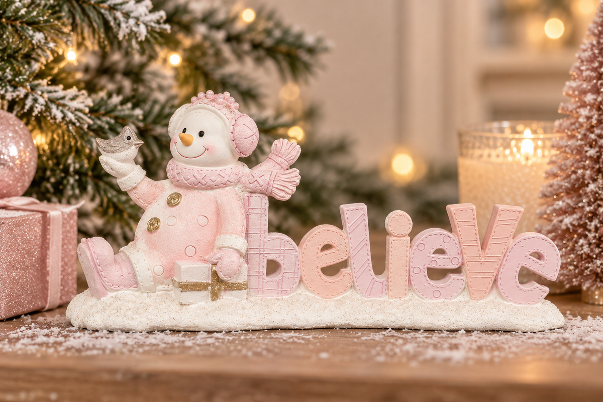 Pink snowman 'Believe' sign figurine displayed on a wooden table with a pink gift box and blush Christmas tree in warm bokeh light