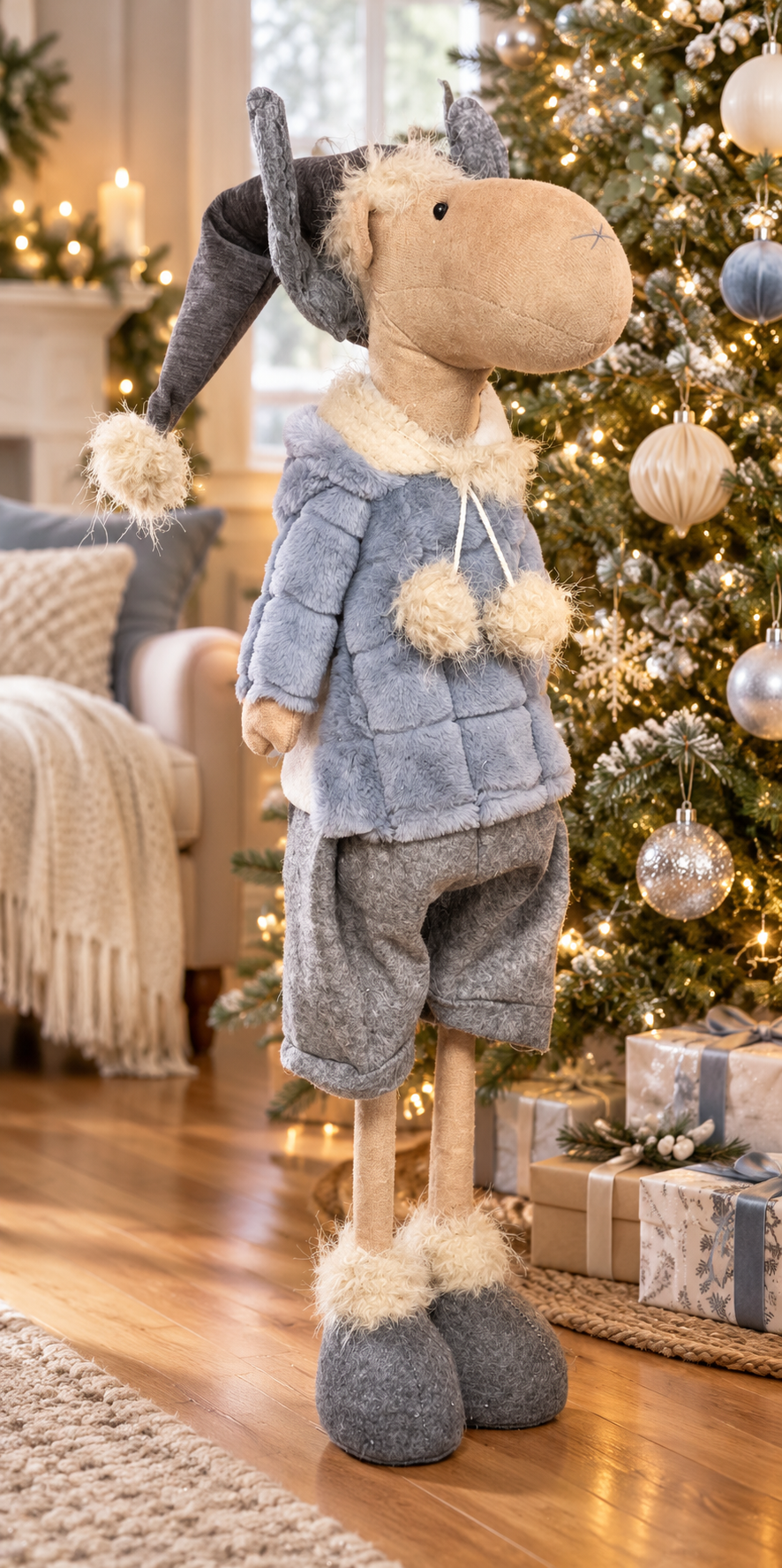 Tall standing Christmas moose in a blue quilted faux fur jacket, grey shorts and a grey beanie with pom pom, standing on a timber floor beside a decorated Christmas tree with silver and cream baubles, wrapped gifts and a cosy lounge in the background
