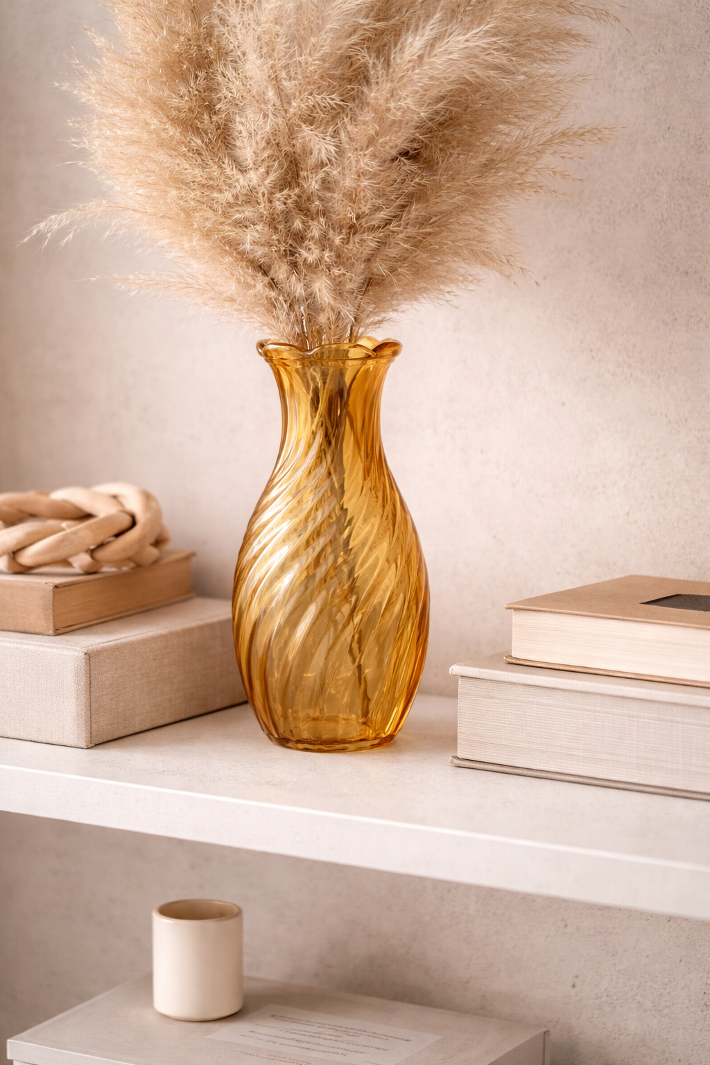 Amber twisted swirl glass vase with pampas grass on a white shelf styled with stacked books and a small candle — Mistletoe Home