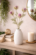 Faux purple thistle spray stems arranged in a white textured vase on a timber console table styled with stacked books, a woven tassel garland, a rattan ball and a pillar candle — Mistletoe Home