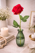 Single faux red rose stem with green leaves in a small green ribbed glass bud vase styled on a neutral table with a white pillar candle, stacked books, wooden bead garland and white gypsophila — Mistletoe Home