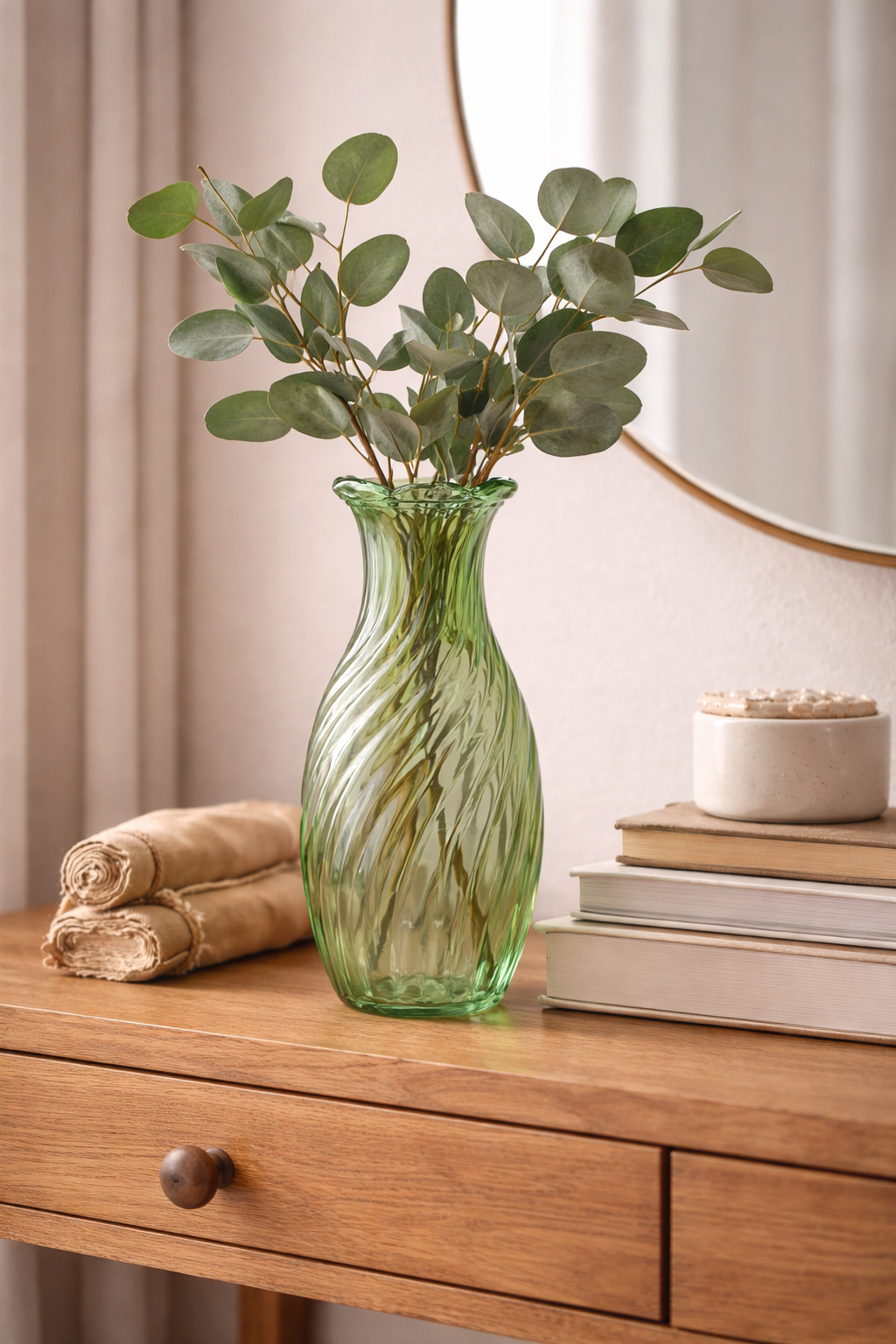 Green twisted swirl glass vase with eucalyptus stems on a timber dresser beside stacked books and a round mirror — Mistletoe Home