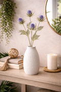 Faux blue thistle spray stems arranged in a white textured vase on a timber console table styled with stacked books, a woven tassel garland, a rattan ball and a pillar candle — Mistletoe Home