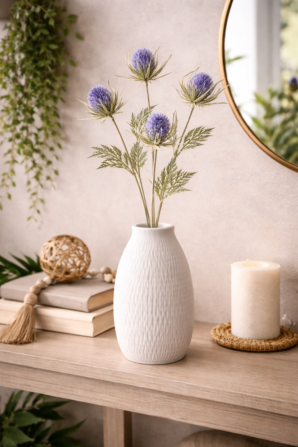 Faux blue thistle spray stems arranged in a white textured vase on a timber console table styled with stacked books, a woven tassel garland, a rattan ball and a pillar candle — Mistletoe Home
