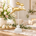 Gold leaping bunny ornament with cream ribbon bow on a white stand, styled on a wooden table with white tulips, pink blossoms, a woven tray of gold and cream Easter eggs and a cream table runner