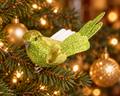 Lime green glitter bird ornament with velvety head and wings clipped to a Christmas tree branch, surrounded by warm fairy lights and gold baubles