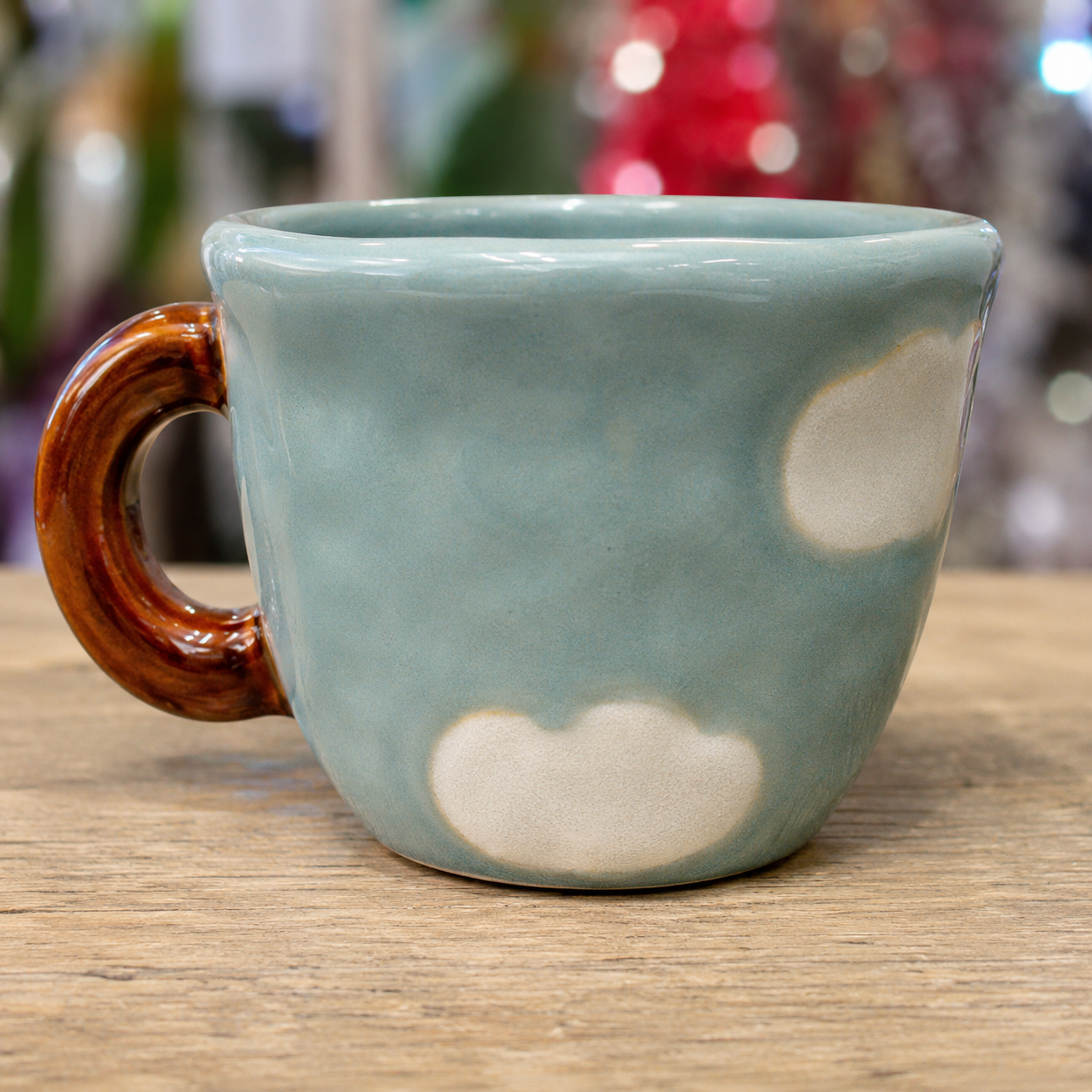 Close-up of Nini Cup Clouds kids' mug on a wooden table with blurred festive bokeh lights in the background, highlighting the cloud motif and brown handle