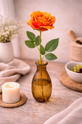Single faux orange rose stem with rich ombré petals and green leaves in an amber ribbed glass bud vase styled on a neutral table with a lit pillar candle on a wicker trivet, white gypsophila and a wooden mortar — Mistletoe Home