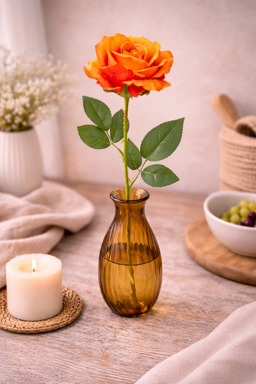 Single faux orange rose stem with rich ombré petals and green leaves in an amber ribbed glass bud vase styled on a neutral table with a lit pillar candle on a wicker trivet, white gypsophila and a wooden mortar — Mistletoe Home