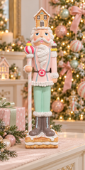 Pastel gingerbread nutcracker figurine with a gingerbread house hat, white icing beard, pink jacket, mint trousers and candy cane staff, standing on a biscuit base on a cream sideboard beside a blush and mint Christmas tree and a pink wrapped gift