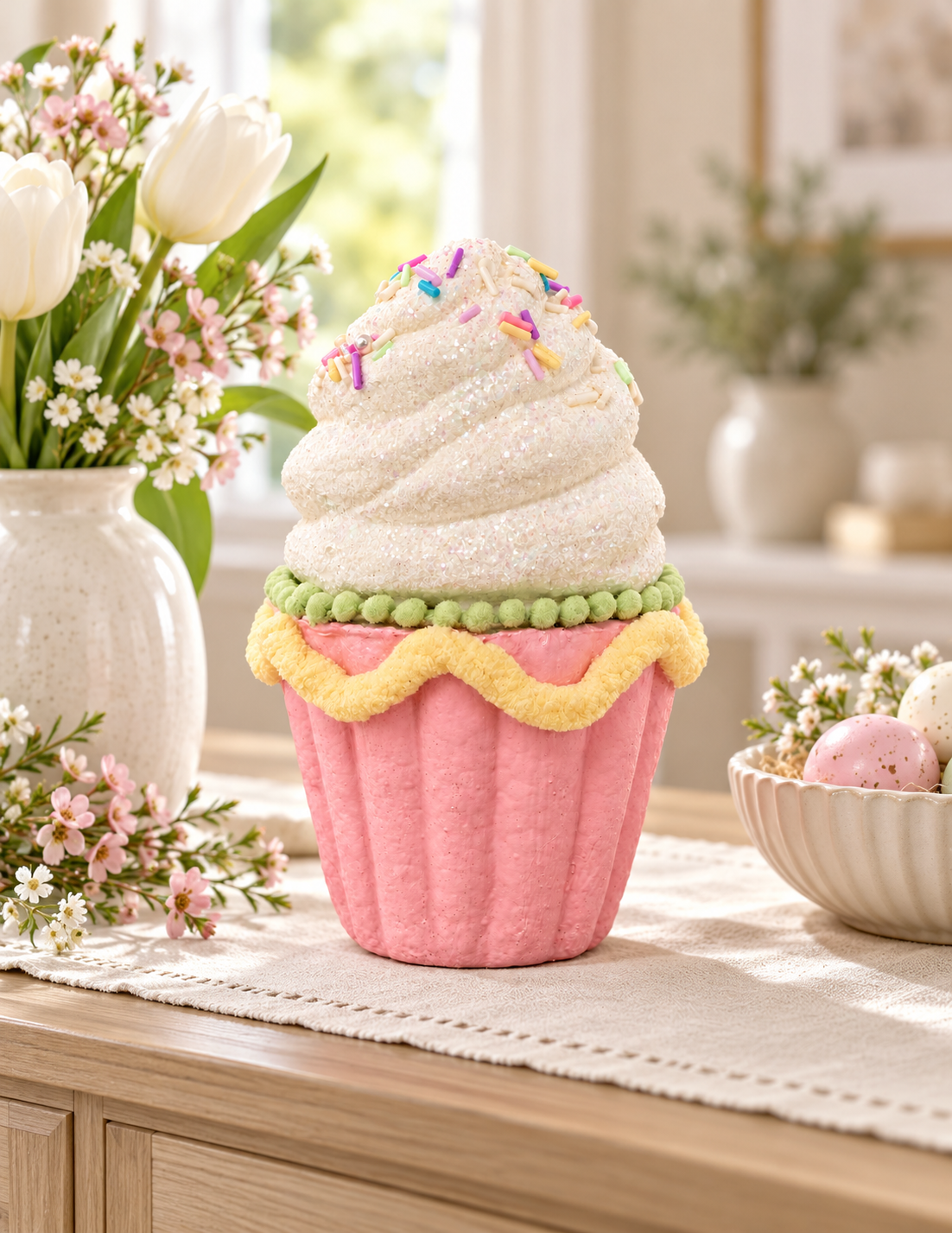Pink sprinkle cupcake decoration with white glitter frosting and rainbow sprinkles, styled on a timber sideboard with white tulips, pink blossoms and a bowl of speckled Easter eggs