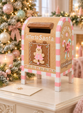 Pink gingerbread Santa mailbox with pink and white candy cane stripes, a gingerbread girl door and “letter to Santa” lettering, styled on a white sideboard with a pink candle holder, an envelope, frosted pine sprigs and a pastel Christmas tree