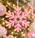 Pink glitter snowflake ornament with white piped icing, gold tips and rainbow sprinkles hanging from a white ribbon on a Christmas tree with pink baubles, a peppermint bauble and a candy cane