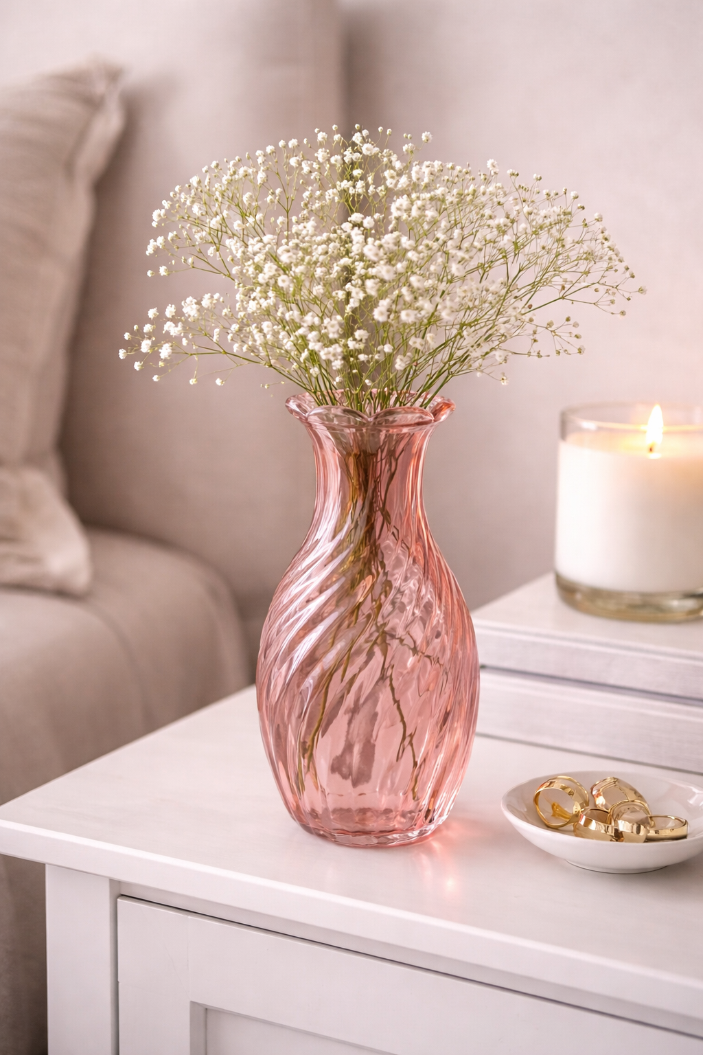 Pink twisted swirl glass vase with white baby's breath flowers on a bedside table beside a lit candle and gold rings — Mistletoe Home