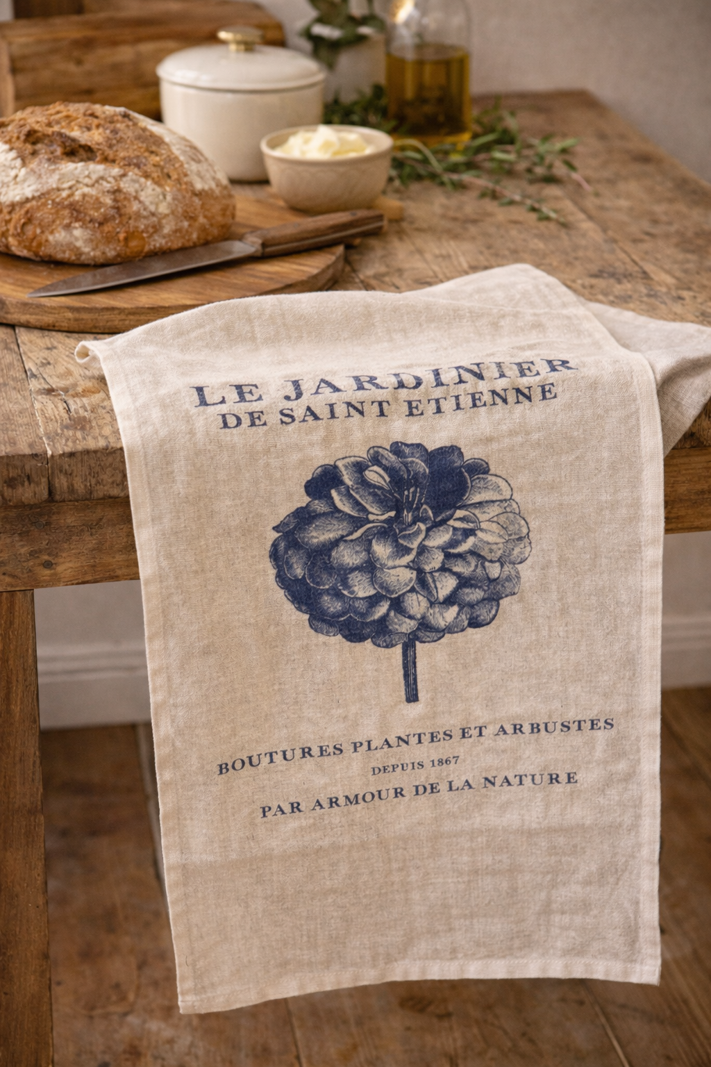 Natural linen tea towel with navy Le Jardinier de Saint Etienne French botanical print draped over a rustic timber kitchen table with sourdough bread, olive oil and fresh herbs — Mistletoe Home
