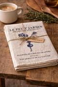 Le Petit Jardin linen tea towel folded and tied with natural twine bow and 100% linen tag, styled on a rustic timber table with coffee cup, wooden spoon and fresh thyme — Mistletoe Home