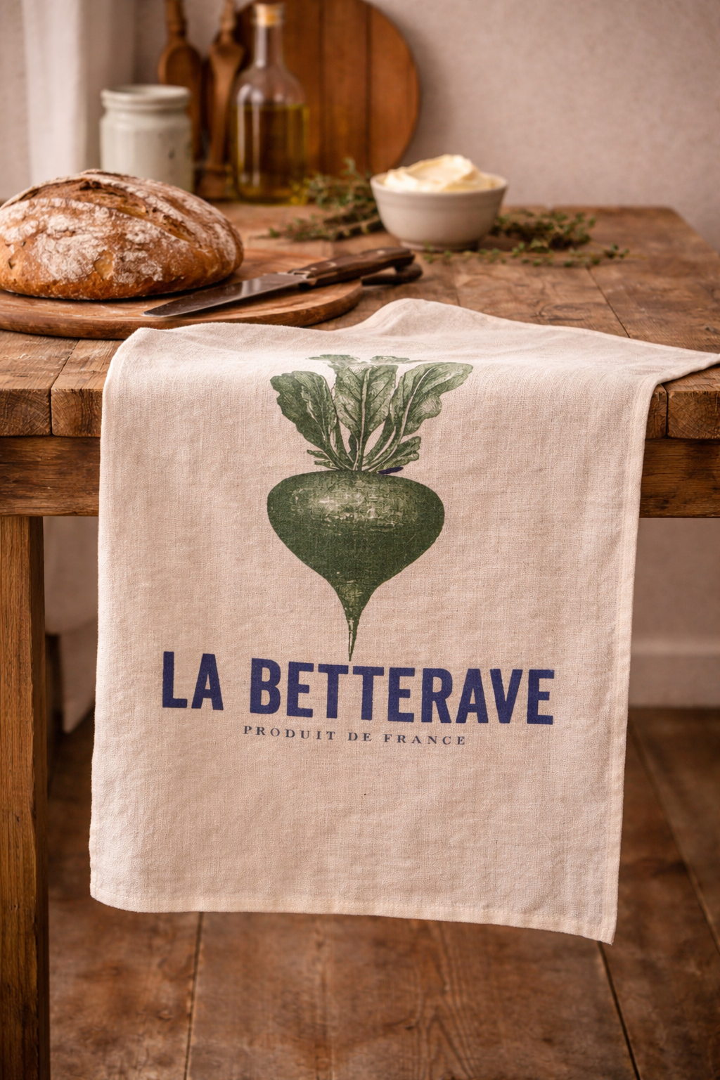Natural linen tea towel with navy and green La Betterave French beetroot botanical print draped over a rustic timber kitchen table with sourdough bread, olive oil and fresh herbs — Mistletoe Home
