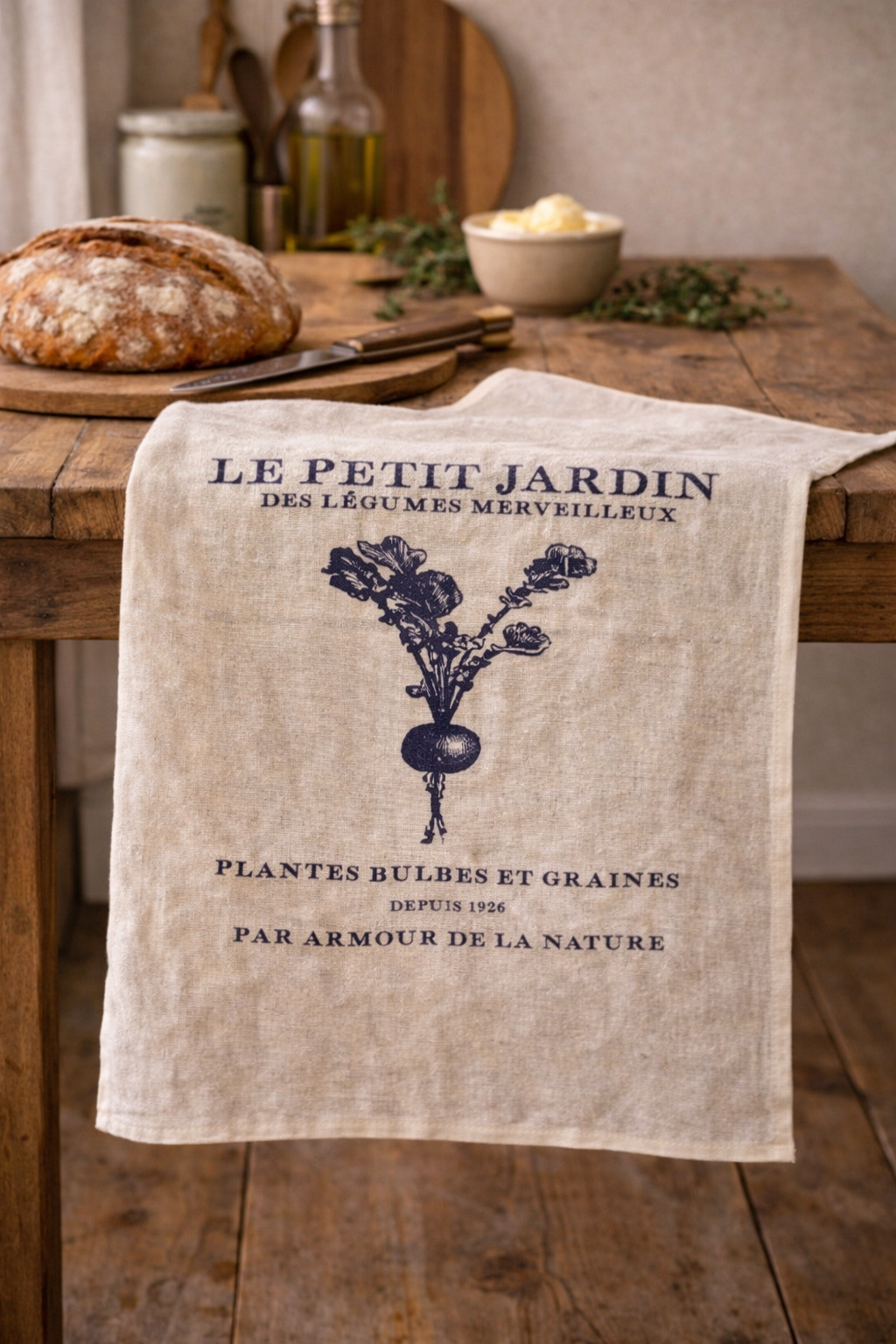  Natural linen tea towel with navy Le Petit Jardin French botanical garden print draped over a rustic timber kitchen table with sourdough bread, olive oil and fresh herbs — Mistletoe Home