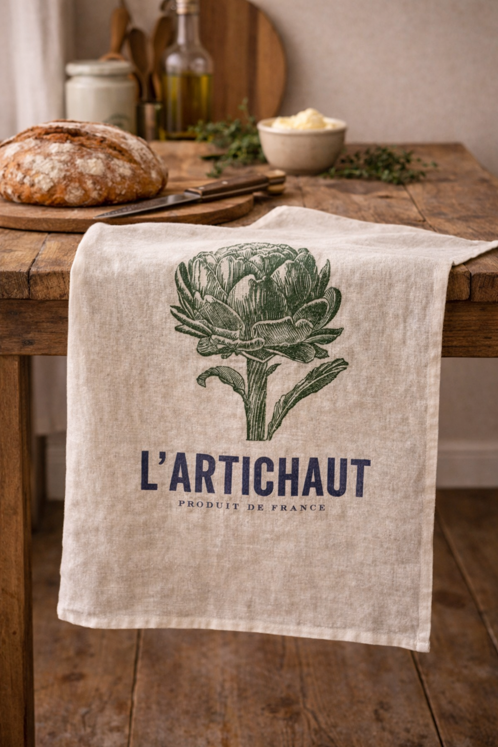 Natural linen tea towel with navy and green L'Artichaut French artichoke botanical print draped over a rustic timber kitchen table with sourdough bread, olive oil and fresh herbs — Mistletoe Home