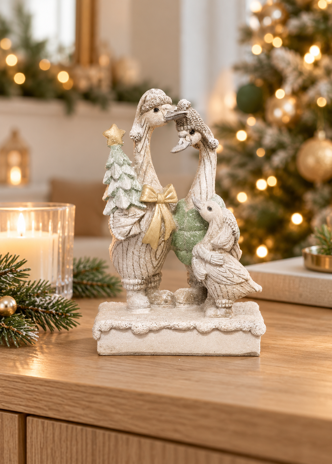 Christmas goose family figurine with two geese in knit beanies sharing a kiss, a gosling in a scarf, a mint Christmas tree and gold bow on a snowy base, styled on a wooden sideboard with a candle, pine sprigs and a glowing Christmas tree