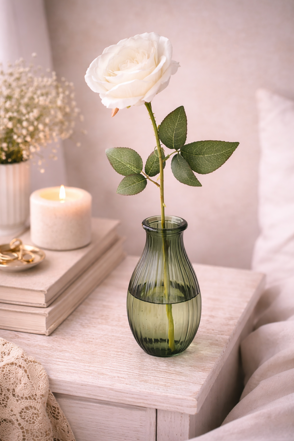 Single faux white rose stem in a small green ribbed glass bud vase on a white timber bedside table styled with stacked books, a lit pillar candle, gold jewellery dish and white gypsophila — Mistletoe Home