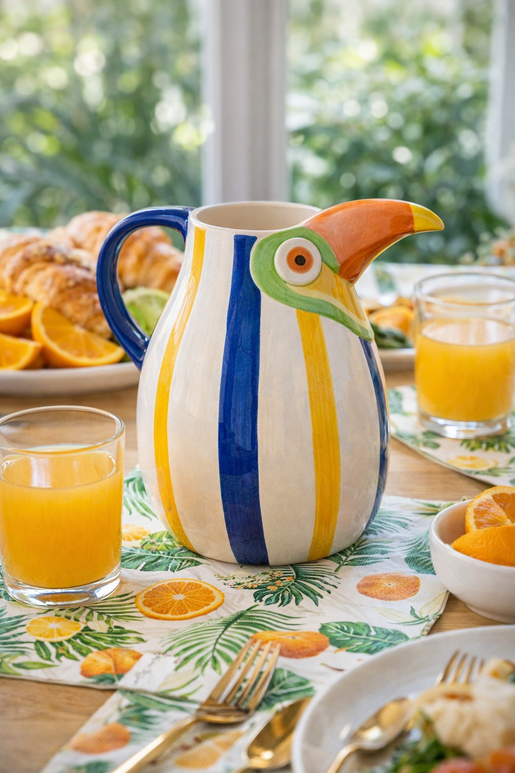 Large hand-painted ceramic toucan jug in blue, yellow and white stripes with sculptural orange beak, styled on a tropical brunch table with orange juice, croissants and fresh oranges — Mistletoe Home