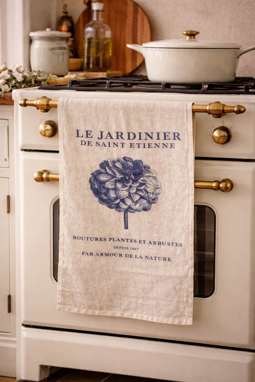 Natural linen Le Jardinier tea towel with navy dahlia botanical print hanging from a cream and brass vintage French oven with white pot on stovetop — Mistletoe Home