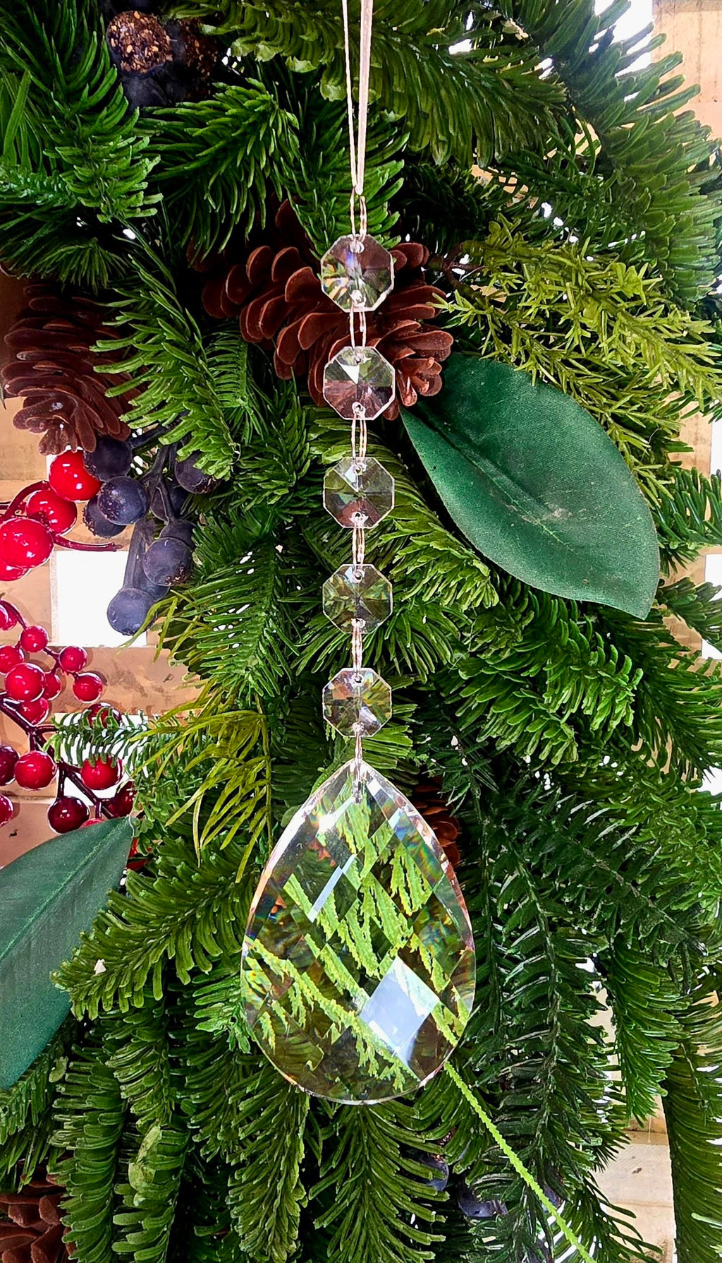 Clear crystal pear drop ornament hanging on a Christmas tree decorated with pine cones, red berries and magnolia leaves, with five octagonal crystal beads on a silver chain above a large teardrop pendant