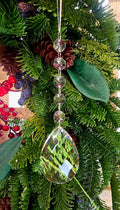 Clear crystal pear drop ornament hanging on a Christmas tree decorated with pine cones, red berries and magnolia leaves, with five octagonal crystal beads on a silver chain above a large teardrop pendant