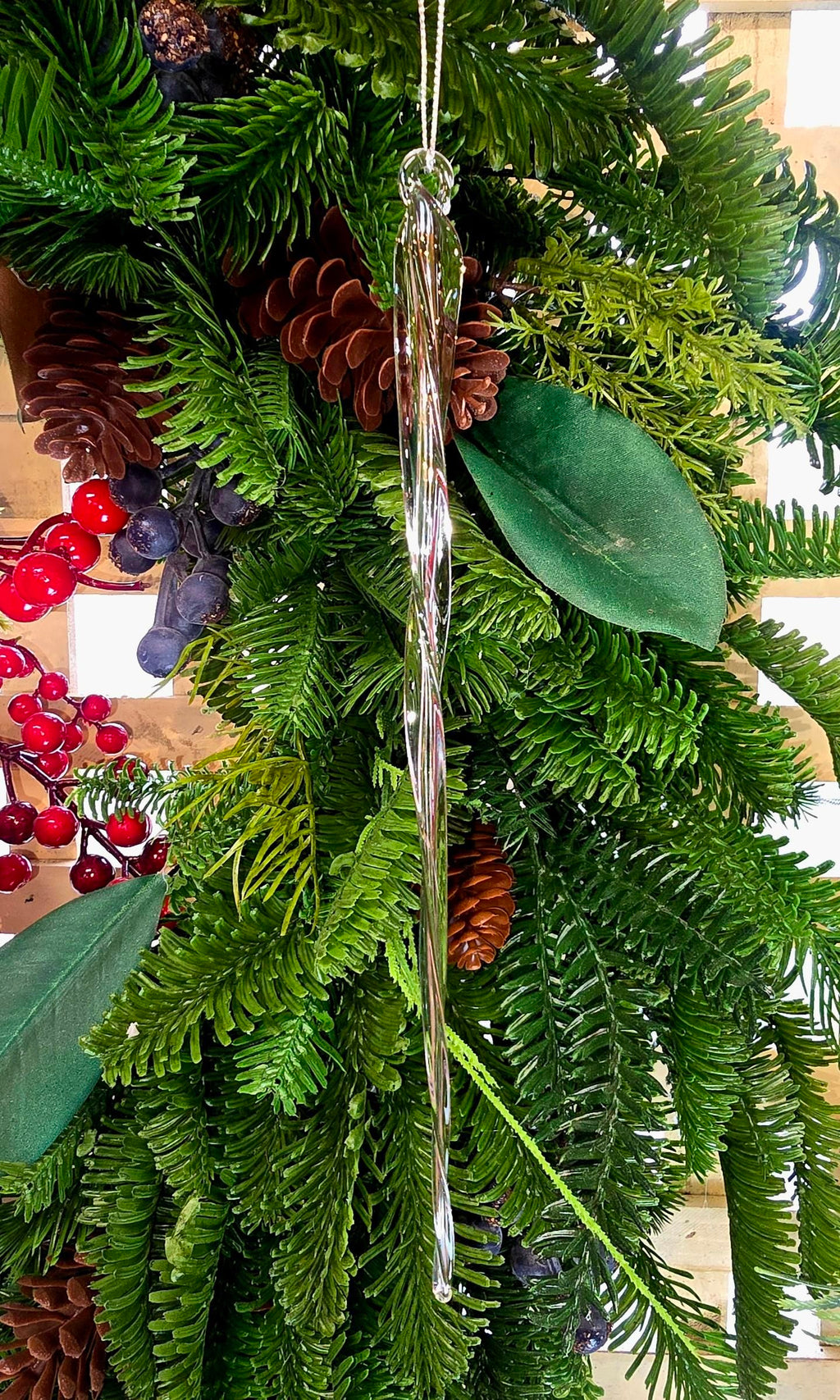 Clear glass braided icicle Christmas ornament hanging on a lush green tree with pine cones, red berries and magnolia leaves, showing the elongated multi-strand twisted form