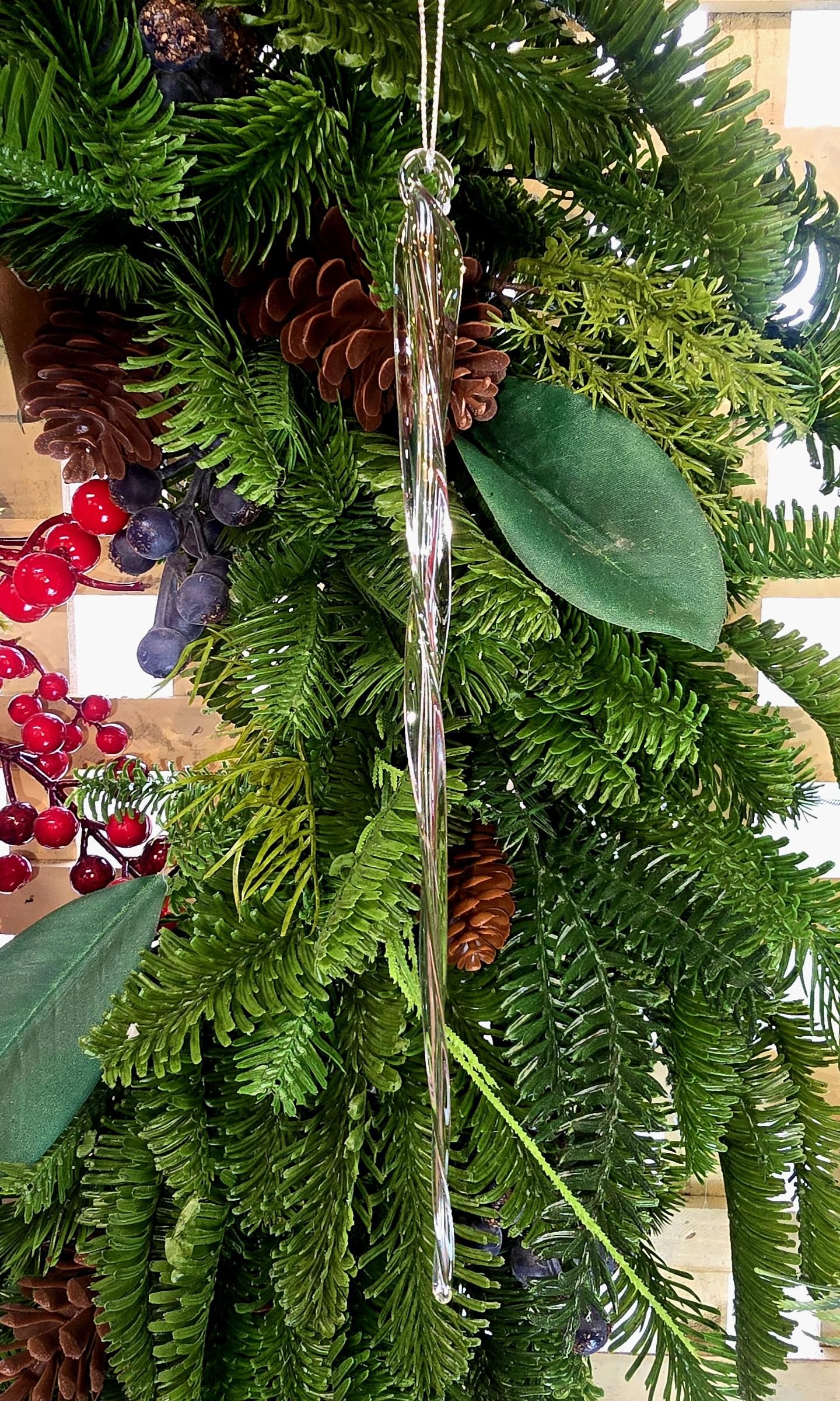 Clear glass braided icicle Christmas ornament hanging on a lush green tree with pine cones, red berries and magnolia leaves, showing the elongated multi-strand twisted form