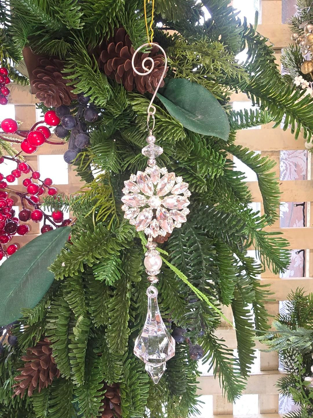 Crystal snowflake ornament hanging on a Christmas tree decorated with pine cones, red berries and magnolia leaves, showing the pearl bead detail and faceted crystal finial drop from the front