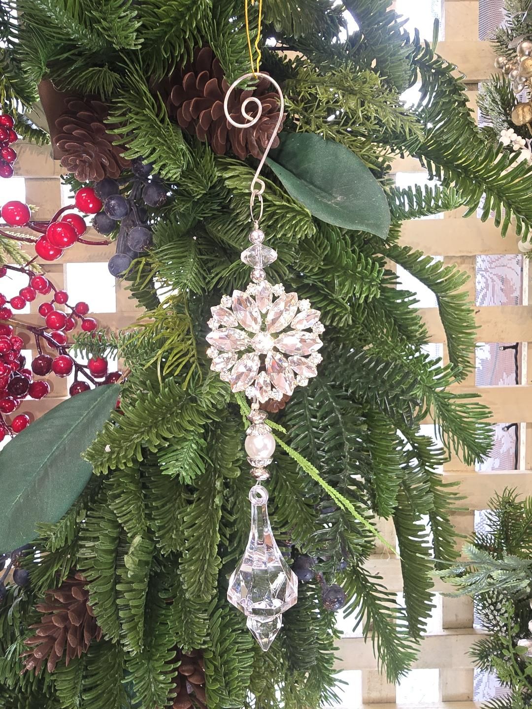 Crystal snowflake ornament hanging on a Christmas tree decorated with pine cones, red berries and magnolia leaves, showing the pearl bead detail and faceted crystal finial drop from the front