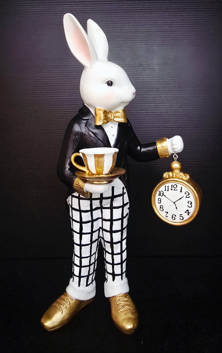 Decorative figurine of a rabbit in formal attire holding a tea cup and pocket watch against a dark background.