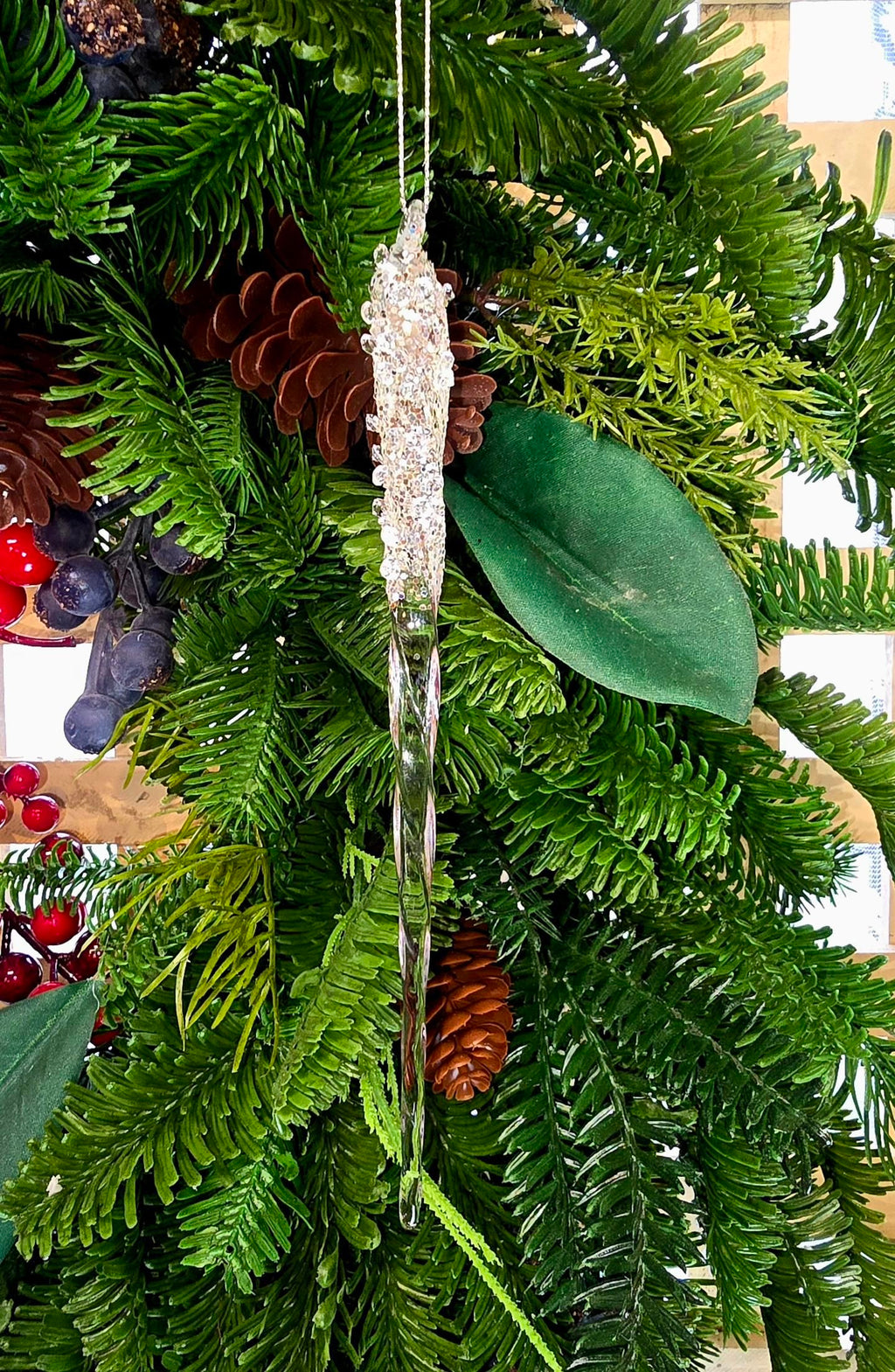 Close-up of the Encrusted Icicle ornament nestled in a pine and pinecone wreath, showing the spiral-twisted glass body and sparkling crystal-bead top