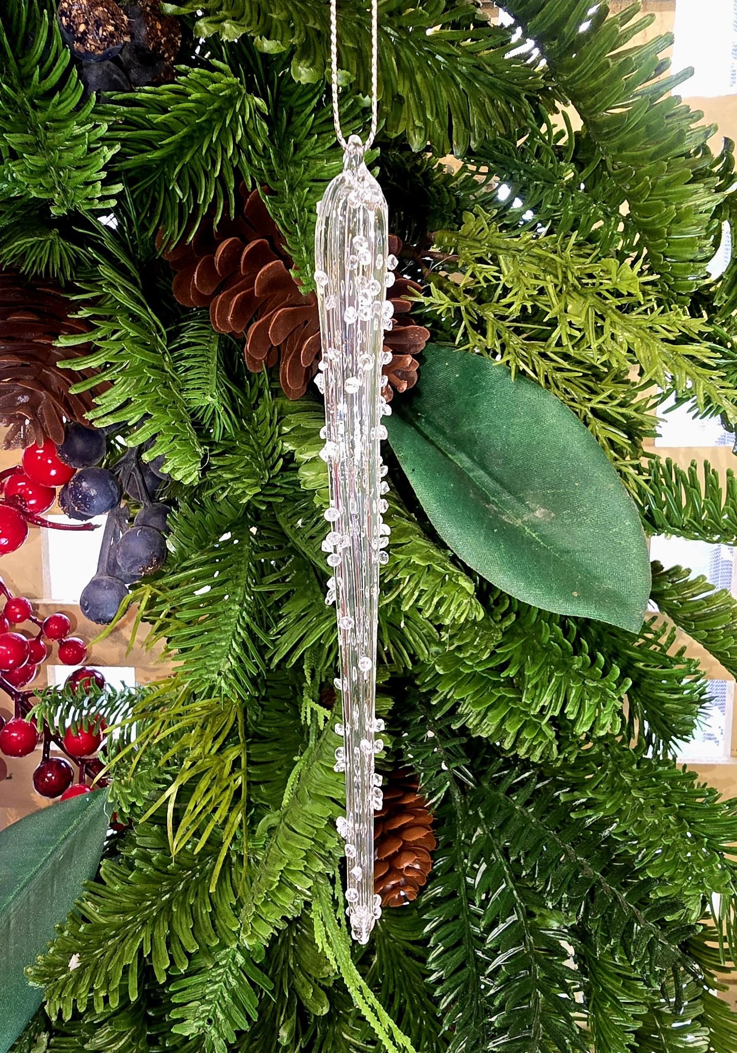 Encrusted glass icicle ornament hanging against a green wreath with pinecones, berries, and magnolia leaves
