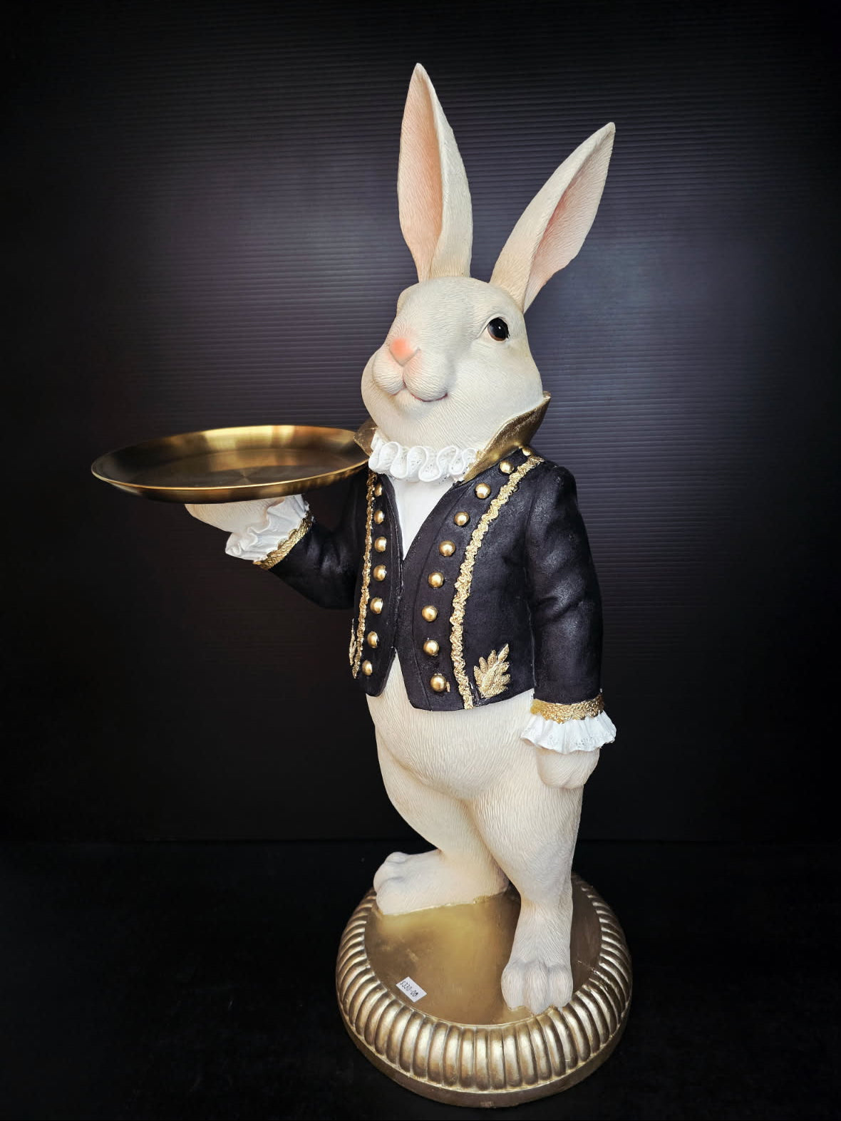 Statue of a rabbit in a formal outfit holding a gold tray against a black background