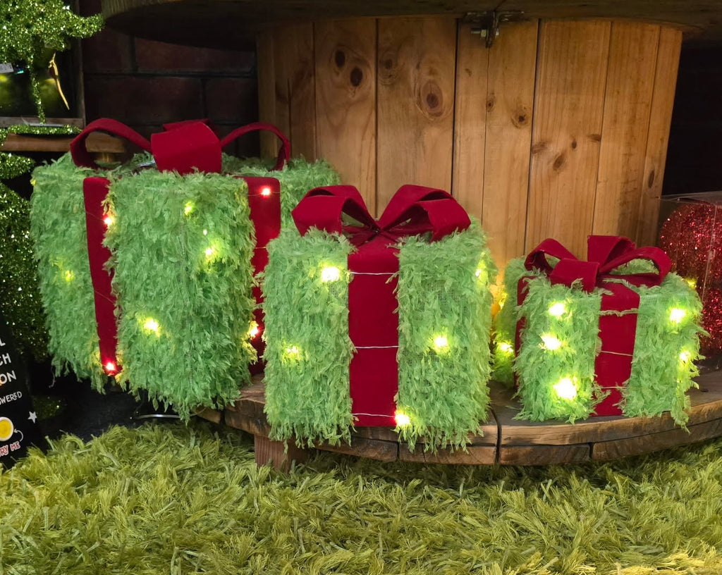 Decorative Christmas lights shaped like gift boxes and holly leaves on a wooden base outdoors.