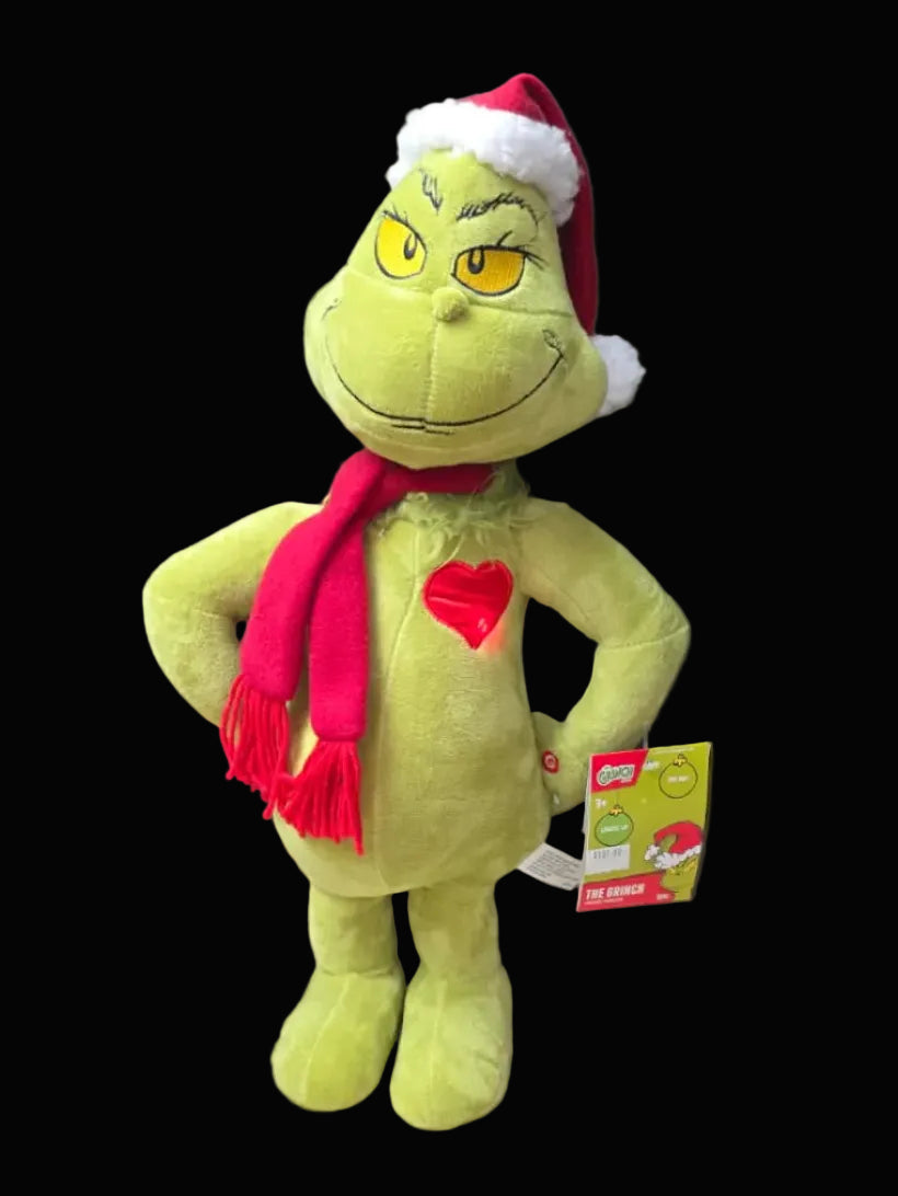 Plush toy resembling a green character with a red heart, Santa hat, and scarf on a dark background.
